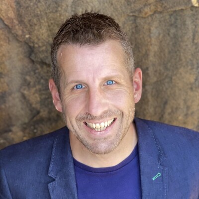 Ben Popplestone Image Smiling man with blue eyes and short brown hair, wearing a blue blazer over a purple shirt, with a rocky wall background.