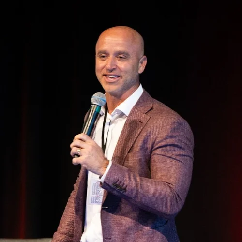 Man in a maroon blazer speaks into a microphone on stage, smiling.