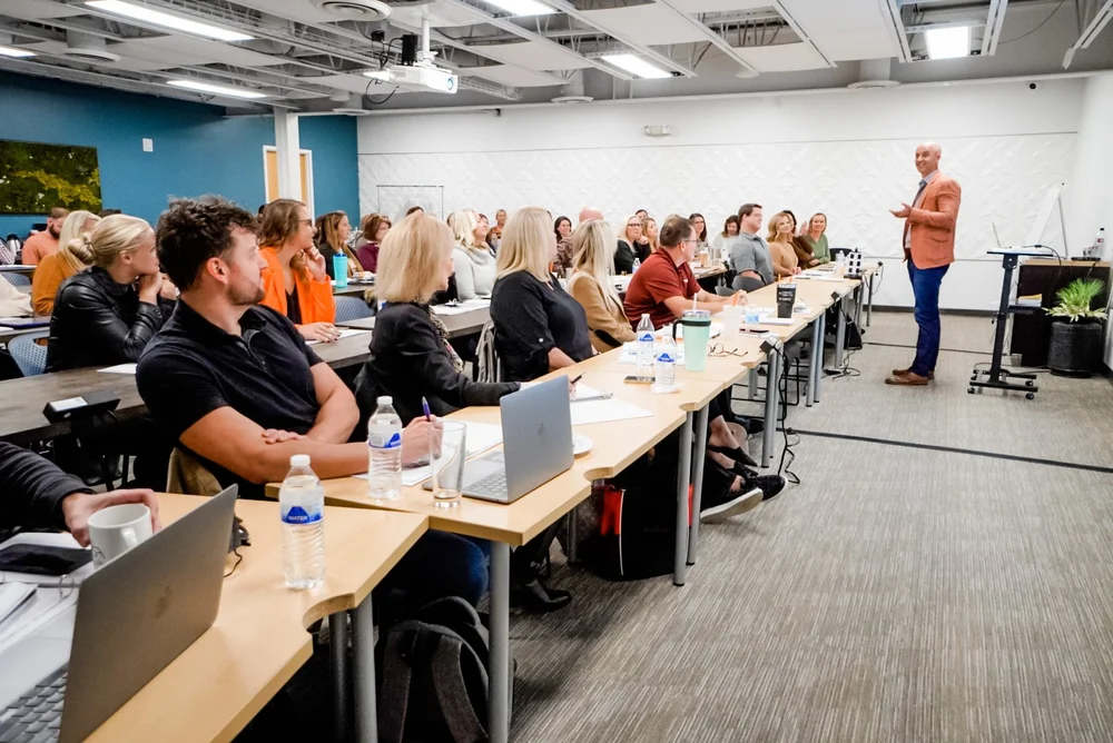 Presenter speaks to a large group seated at long desks in a bright classroom setting.