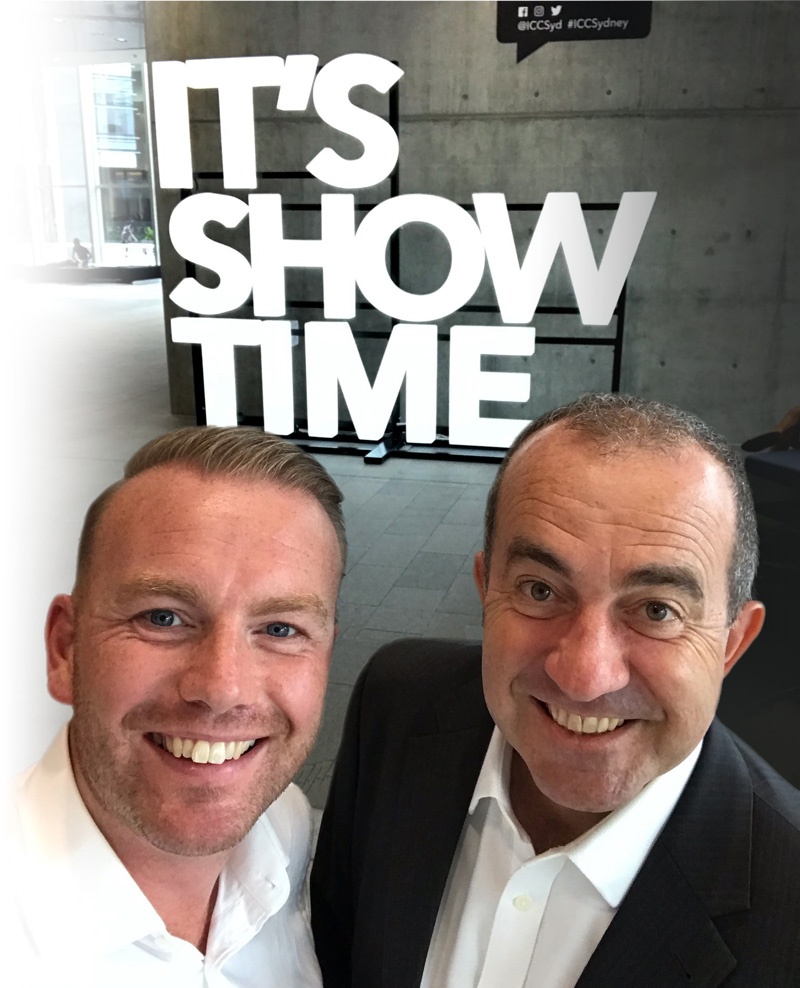 Two men smiling for a selfie in front of a bold 'IT'S SHOWTIME' sign in a modern lobby setting.
